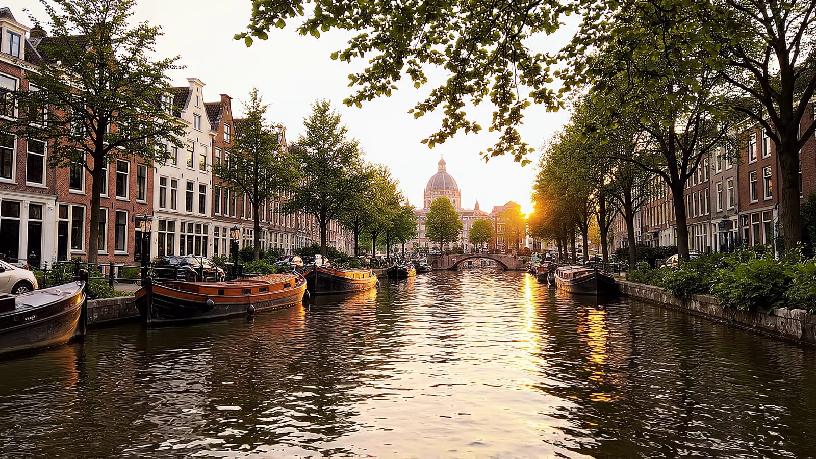 Golden-hour view down an Amsterdam canal: brick gables, leafy trees and calm water catching the last light